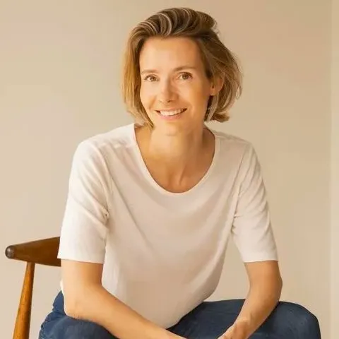 A smiling young woman with medium-length blonde hair wearing a white top sitting in a chair leaning forwards.